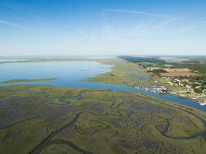 Aerial view looking over a coastal marsh. Ribbons of water curl through the marsh. A small community crowds along the shoreline. Farm fields and forest stretch in the background to the horizon.