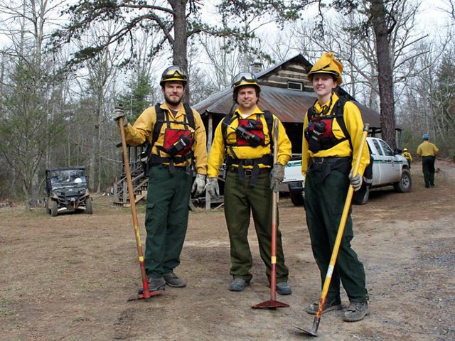 Three people pose together in front of a rustic cabin. They are wearing yellow fire retardant gear and holding long handled hoes. A white pickup truck is parked behind them.