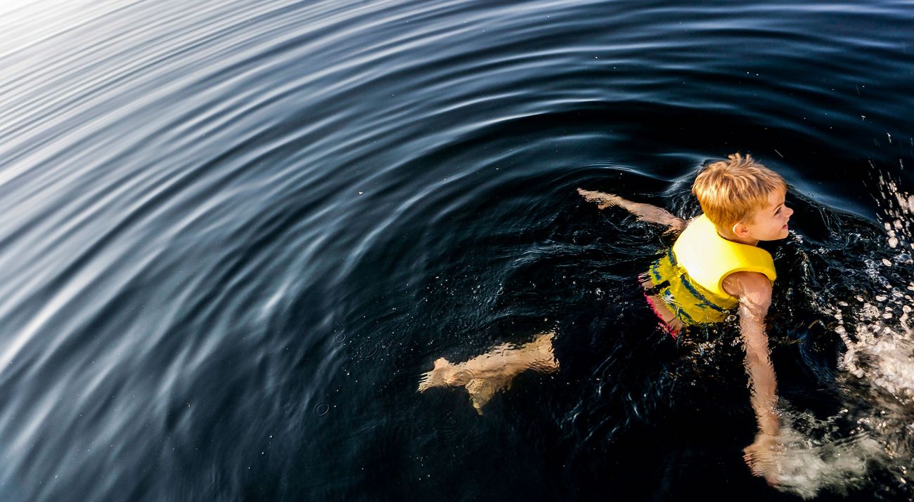 A boy swims in calm, dark blue waters with a yellow life jacket.