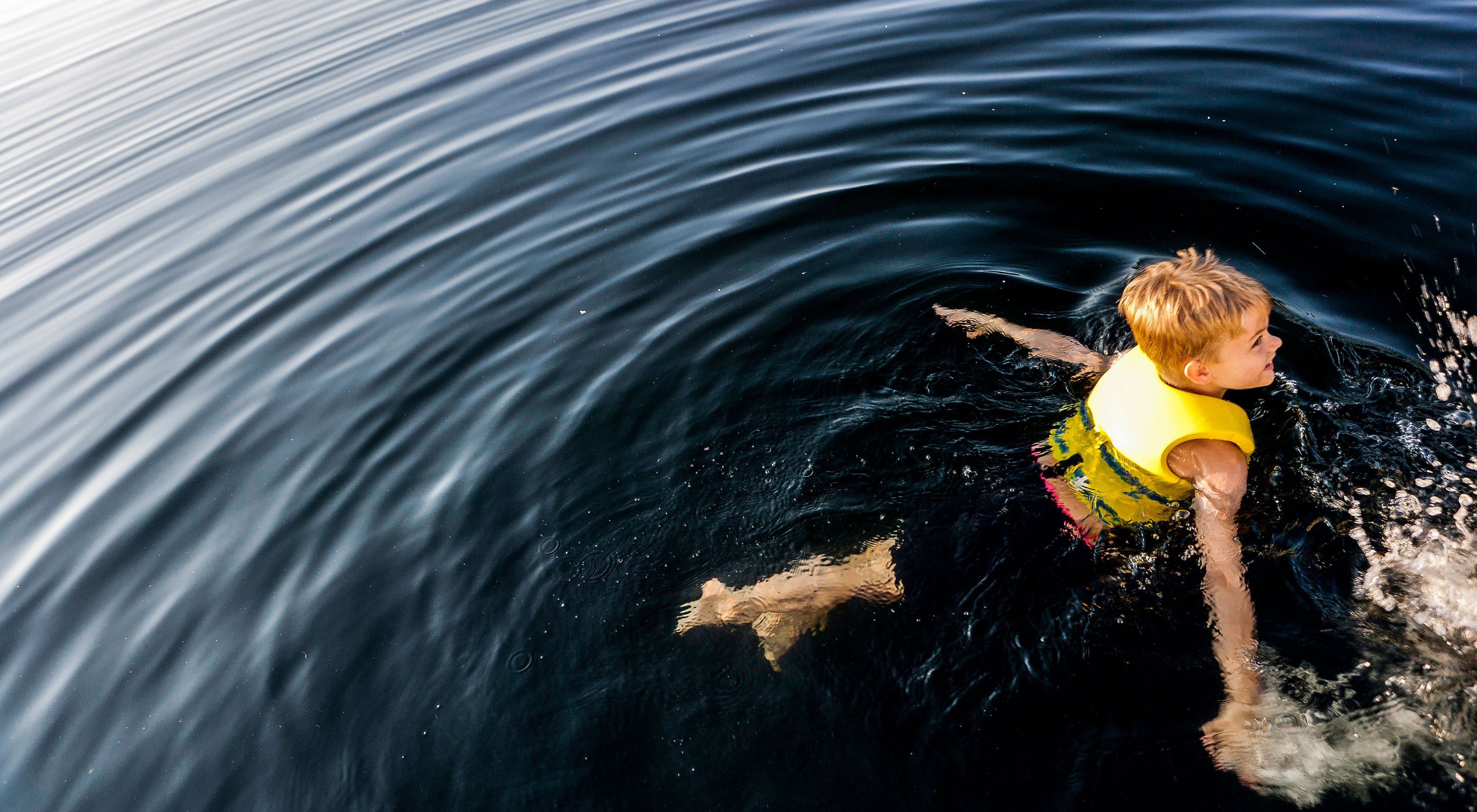 A boy swims in calm, dark blue waters with a yellow life jacket.