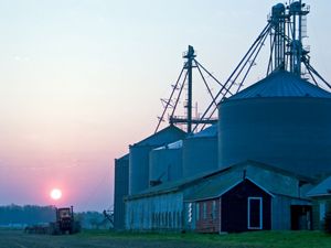 The sun rises over a farm in Maryland. A tractor drives past several low outbuildings flanked by five towering silos.