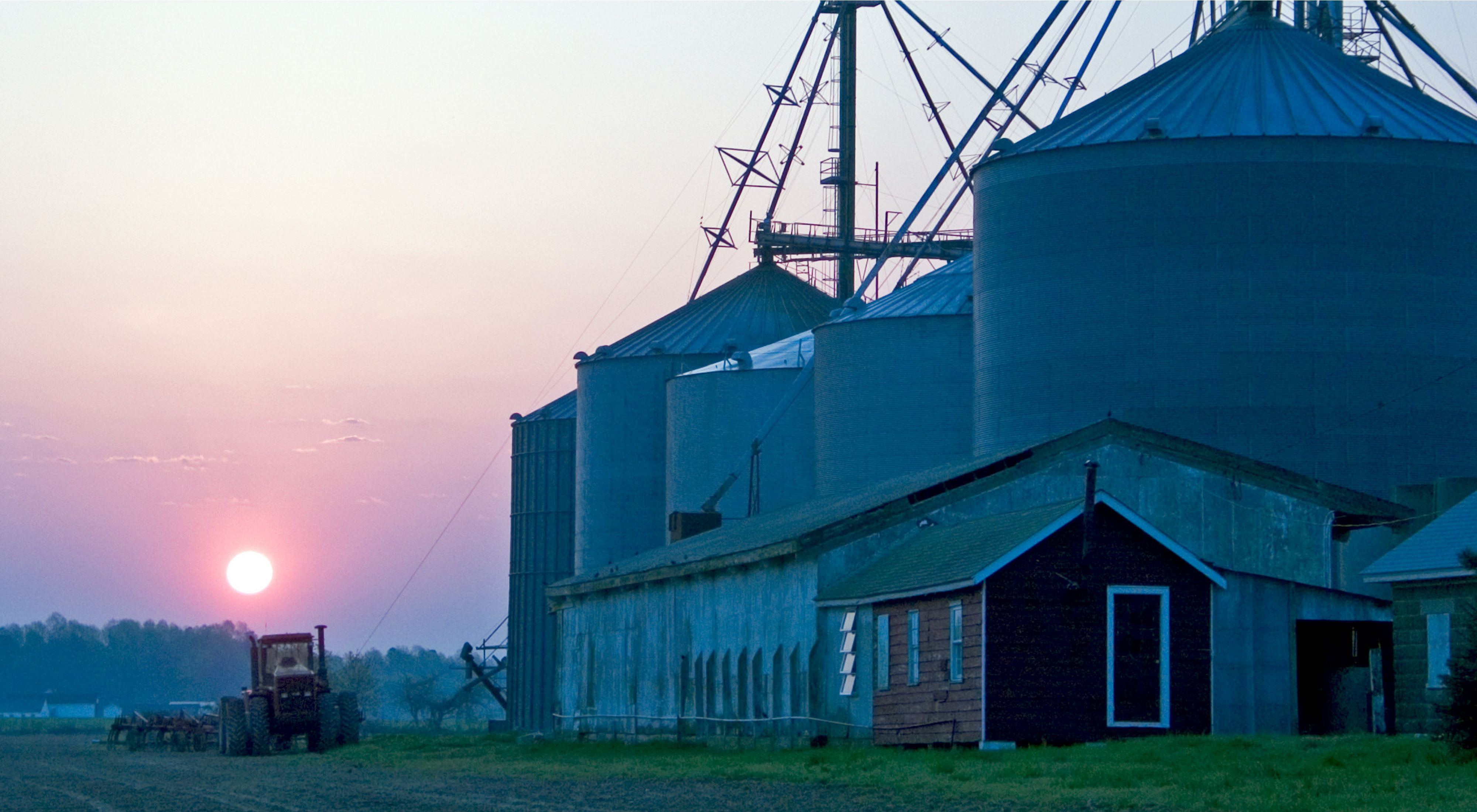 The sun rises over a farm in Maryland. A tractor drives past several low outbuildings flanked by five towering silos.