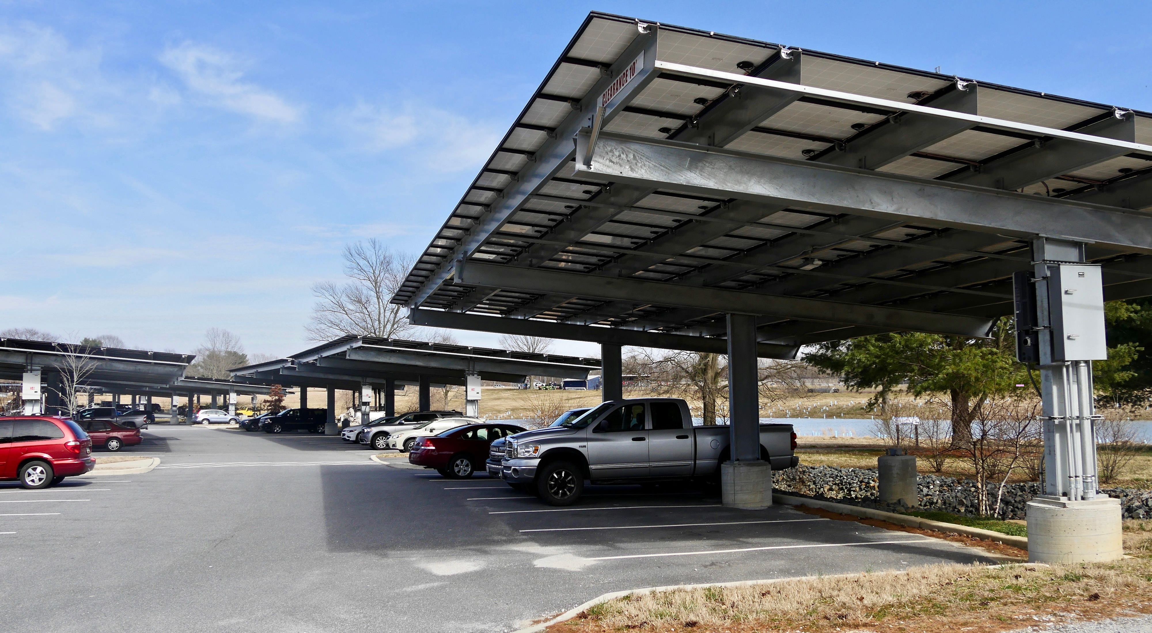 Car and trucks are parked in rows at a commuter parking lot. Large solar arrays spanning 6 or 7 parking spaces shade the vehicles while generating energy.
