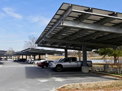 Cars at a commuter lot are parked underneath elevated solar panels that generate energy while shading the vehicles.