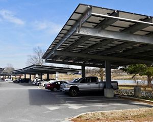 Car and trucks are parked in rows at a commuter parking lot. Large solar arrays spanning 6 or 7 parking spaces shade the vehicles while generating energy.