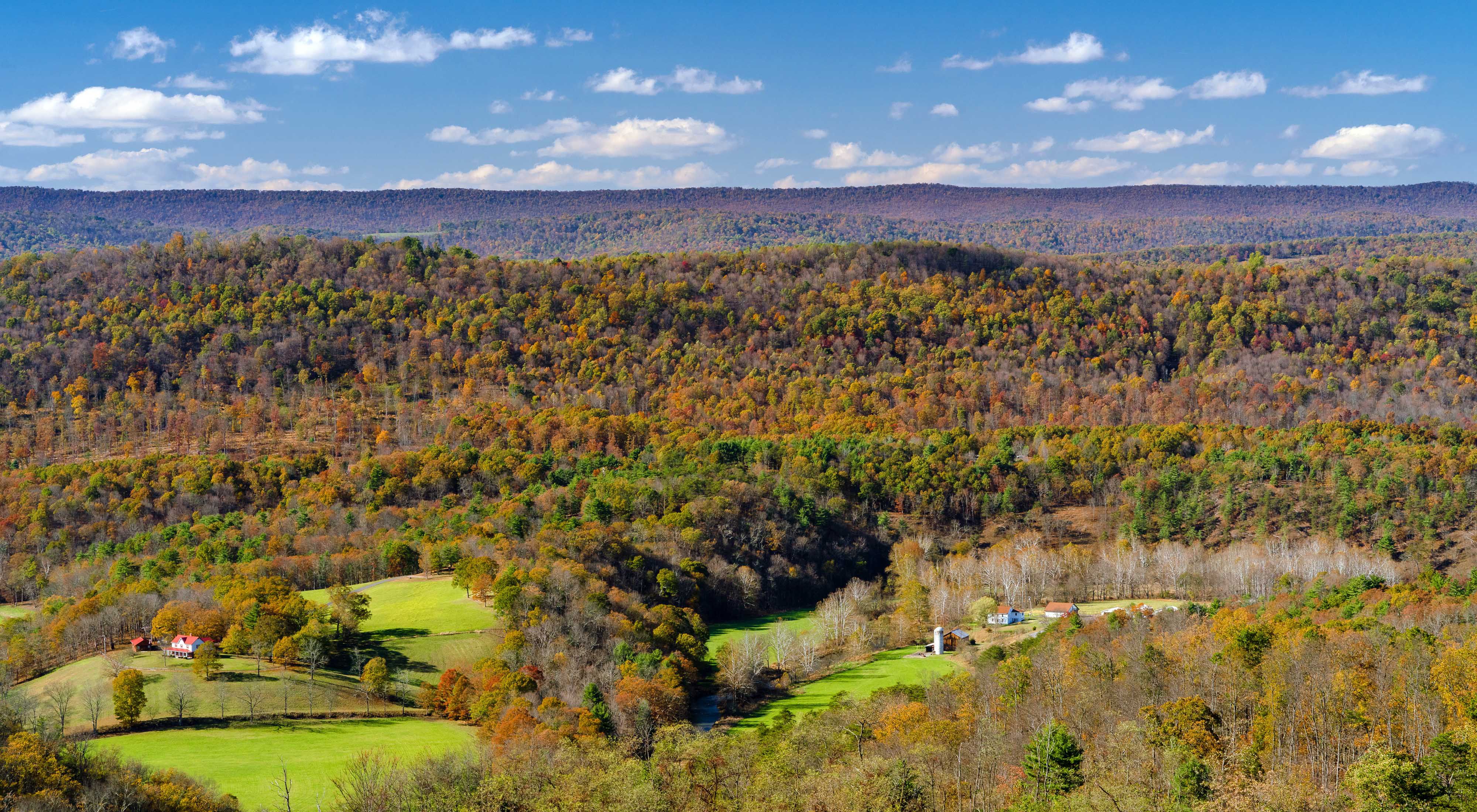 view from elevation looking over a fall deciduous landscape dotted with small farms and gentle, forested mountains in the background under a blue sky