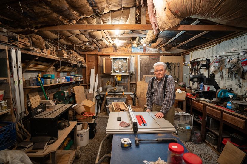 A man standing in a wood shop with wood and tools surrounding him. 