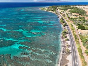 An aerial view of the Honoapi’ilani Highway running on the shore alongside the teal, turquoise, and deep blue colors of the Olowalu Reef, visible under the water from above.