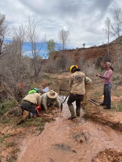 Four people with shovels and other gear work together in a red dirt river.