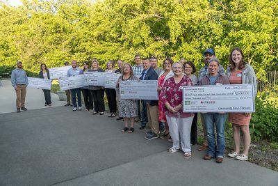 A group of people people pose together outdoors backed by tall trees. Many of the people hold aloft large, novelty checks given out during a grant award ceremony.