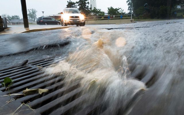 Storm water rushes down a city street into a storm drain. Cars in the background try to navigate the flooded street.
