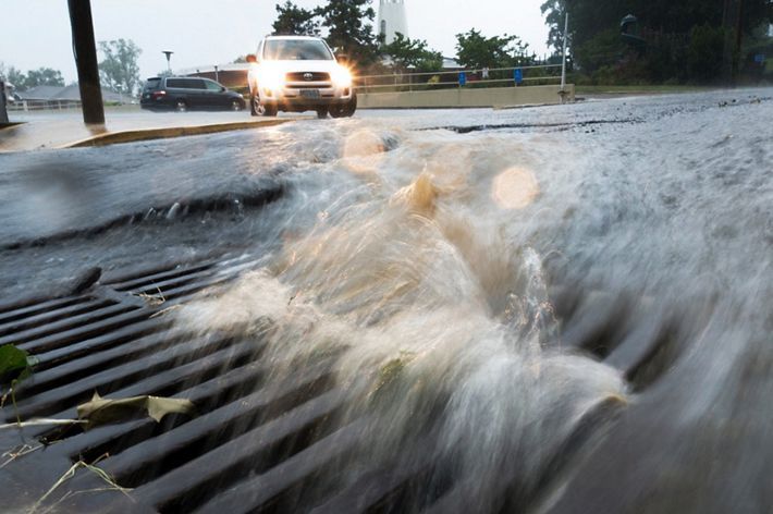 Storm water rushes down a city street into a storm drain. Cars in the background try to navigate the flooded street.