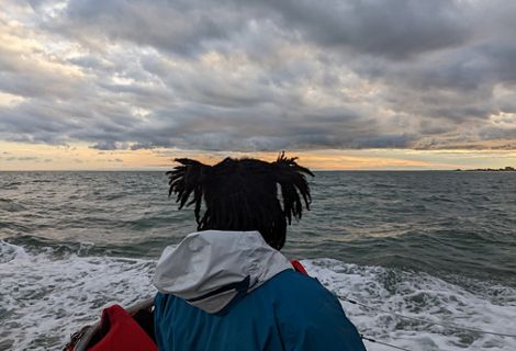 A woman stands on the deck of a boat, facing away from the camera and looking towards the low heavy clouds gathered on the horizon.