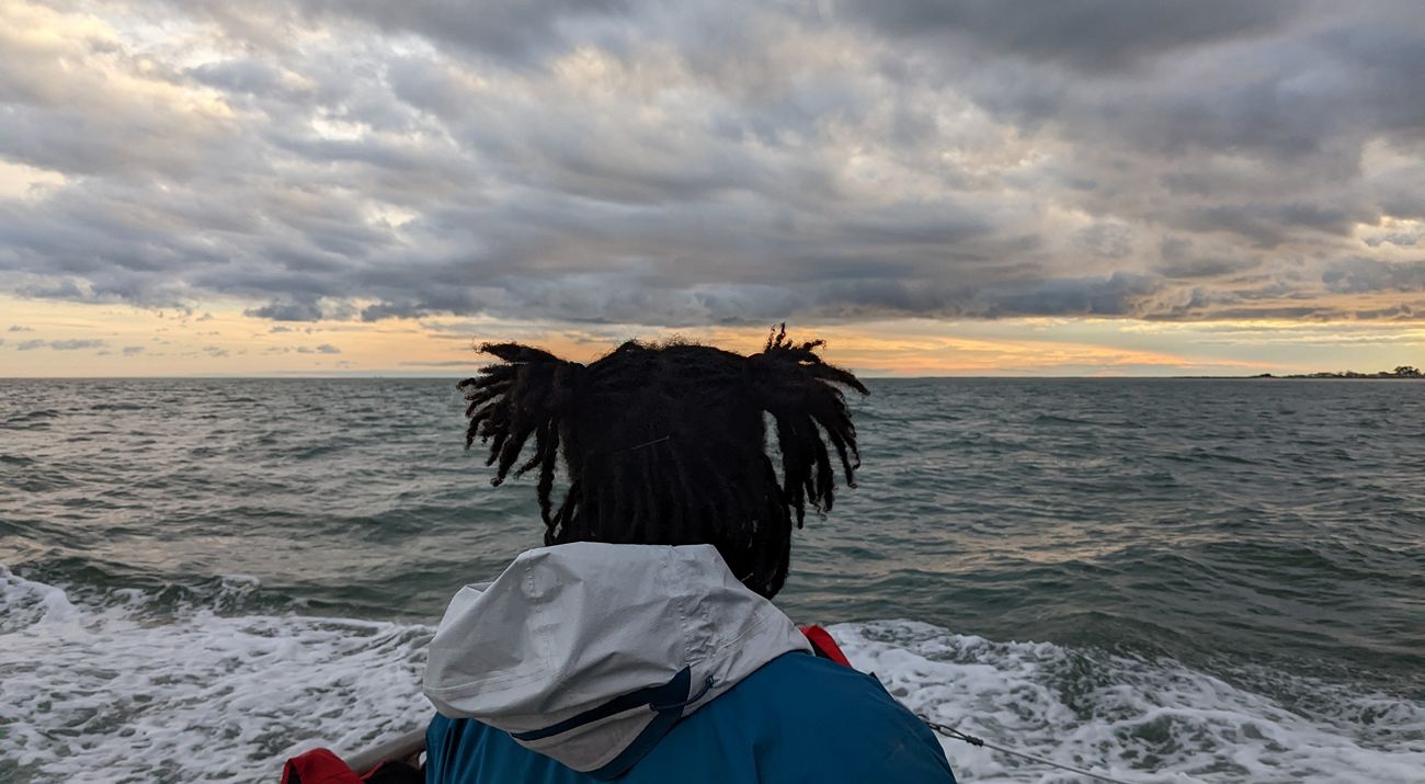A woman stands on the deck of a boat, facing away from the camera and looking towards the low heavy clouds gathered on the horizon.