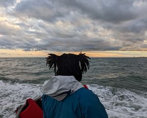 A woman stands on the deck of a boat, facing away from the camera and looking towards the low heavy clouds gathered on the horizon.