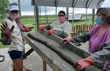 Three women stand around an elevated table using a concrete mixture to create a long, thin piece of oyster reef substrate.
