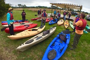 A group of about 10 people stand around two rows of colorful kayaks.