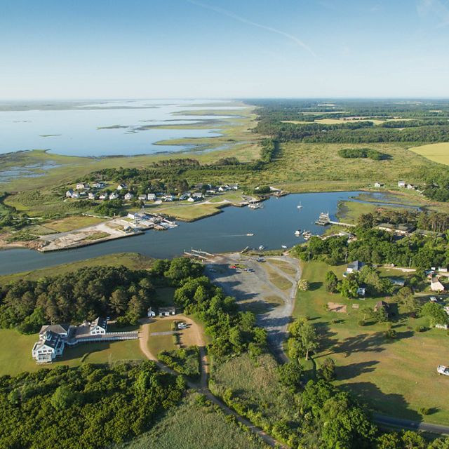 Aerial view of the town of Oyster, VA. Clusters of houses are situated along the edge of a large coastal inlet. Areas of marsh broken up by open water are in the background, extending out to the Atlan