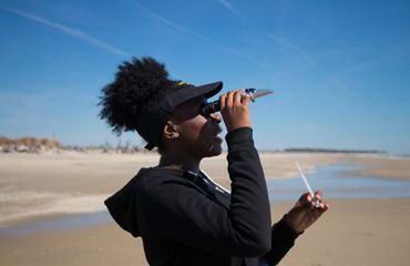 A girl holds a monocular to her eye while conducting science experiments on a beach.