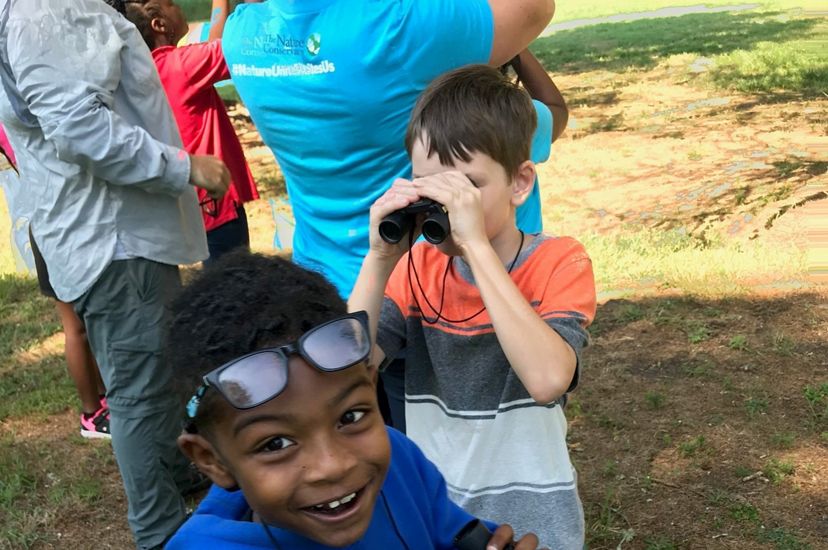 Two boys with binoculars at a summer nature camp. The boy in the background looks through the lenses. The boy in the foreground smiles up at the person taking the photo.