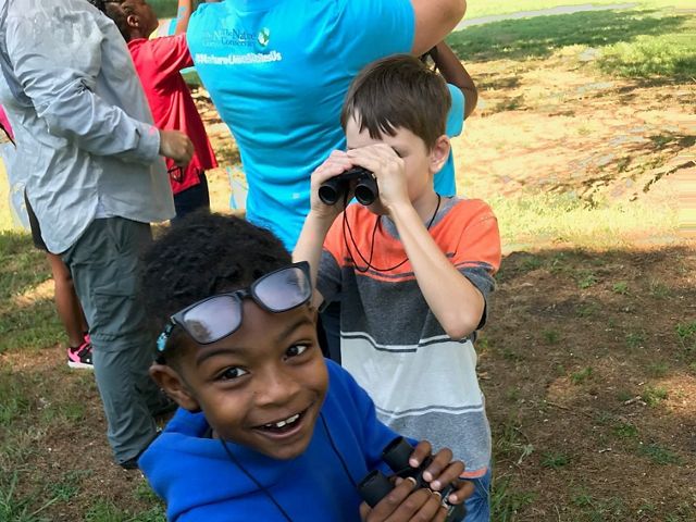 Two boys with binoculars at a summer nature camp. The boy in the background looks through the lenses. The boy in the foreground smiles up at the person taking the photo.