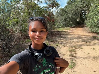 A woman poses for a selfie while she hikes a dirt trail.