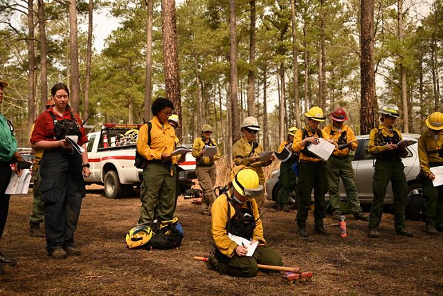 Several men and women in wildland firefighter garb stand in a circle, some taking notes, during their morning pre-fire briefing.