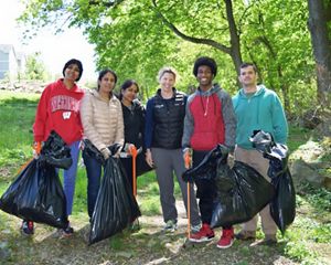 Six people pose together during a stream cleanup event. They are holding large black plastic trash bags and carrying long orange grabbers.