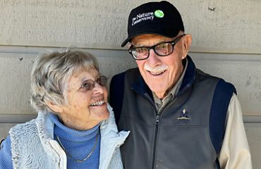 Mary (left) and Vince (right) Ames standing close to each other next to a wooden wall.