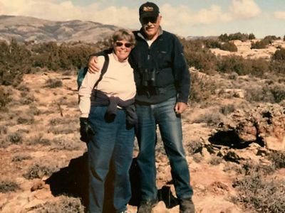 Mary (left) and Vince (right) Ames holding around each other in open landscape with shrubs and a mountain range in the background.