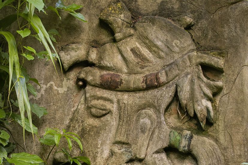 A carved Maya sculpture can be seen through leafy branches.