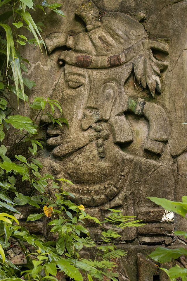 A carved Maya sculpture can be seen through leafy branches.