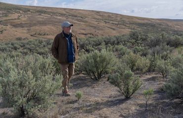 older white man with hat, coat and hiking boots walks among sage green brush with hills stretching behind him
