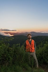 a man in an orange vest stands amid shrubs overlooking mountains in the background during sunset.