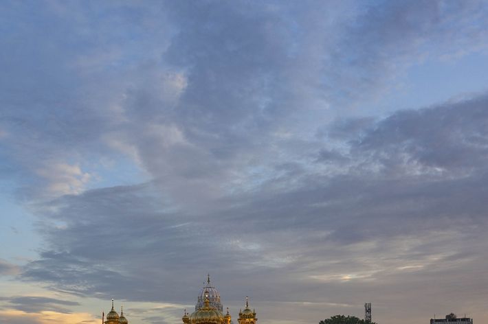 The Golden Temple glows in the twilight and reflects in the water as many people gather on the sidewalk across the river..