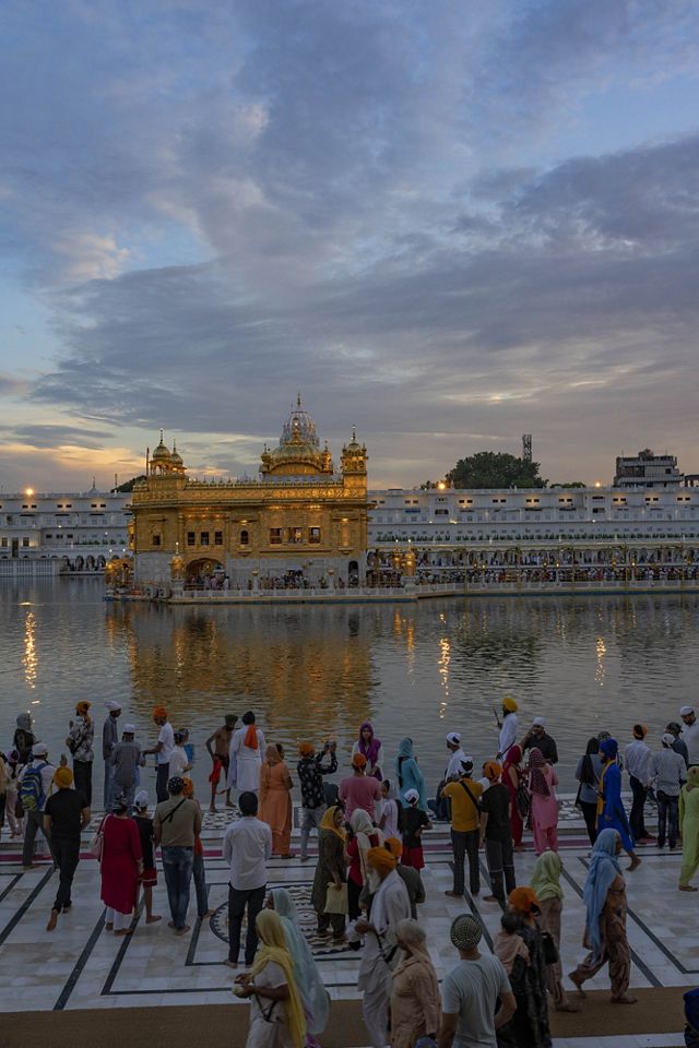 The Golden Temple glows in the twilight and reflects in the water as many people gather on the sidewalk across the river..