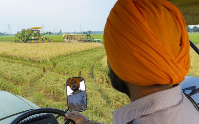 Shot taken over the shoulder of a man driving a tractor in a field; his reflection shows in the tractor's side mirror.