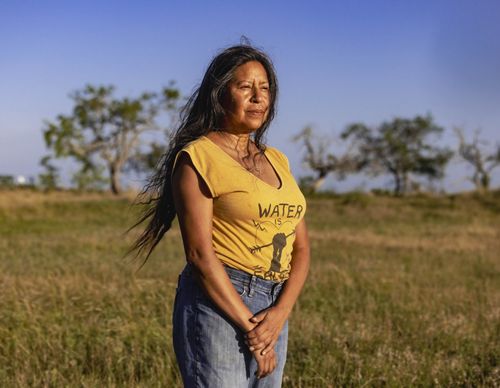 woman stands in yellow shirt, long hair, looking into the distance, brown and green grass and small trees behind her