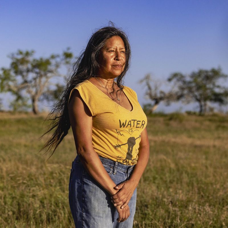 woman stands in yellow shirt, long hair, looking into the distance, brown and green grass and small trees behind her