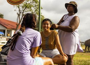 3 women gather: 2 sitting and talking and 1 braiding one of the first women's hair. A horse, barn and other ranch life elements in background