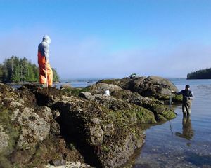 A man in waders stands knee-deep in ocean water while another person wearing waders stands atop a rock jetty.