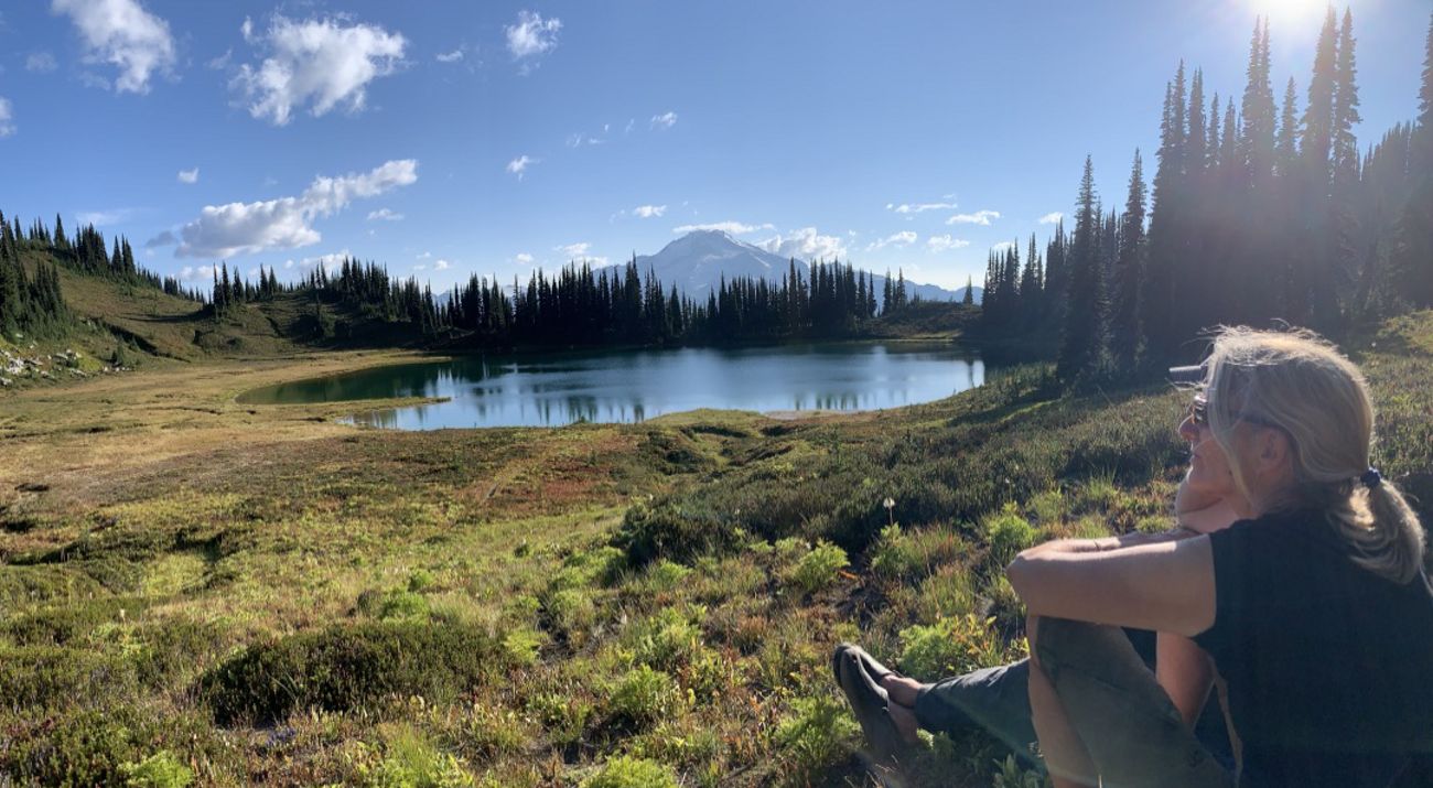 a woman sitting in a prairie with a lake and mountain in the background.