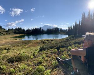 Martha Kongsgaard sits in a grassy meadow with a small lake and a mountain peak in the distance.
