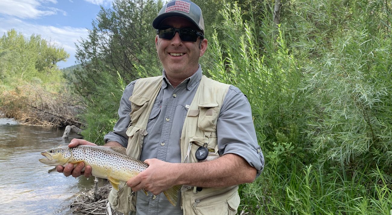 Paul Jewell stands in front of a fresh water stream holding a fish.