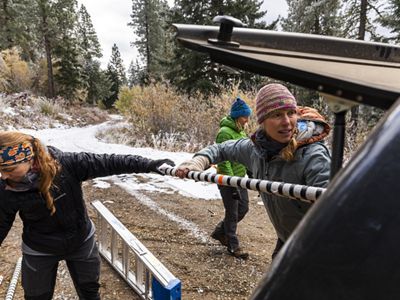 Two people pull equipment out of the back of a vehicle in the middle of a forest.