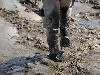 View of the bottom half of a person's legs with tall boots walking through thick mud.