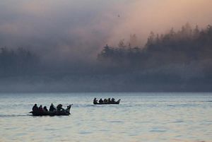 People in two canoes paddle amidst fog toward a forested coastline.