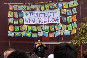 A person stands on a riser addressing a large crowd at a climate march. A collection of hand painted flags hang behind them around a sign reading, protect what you love.