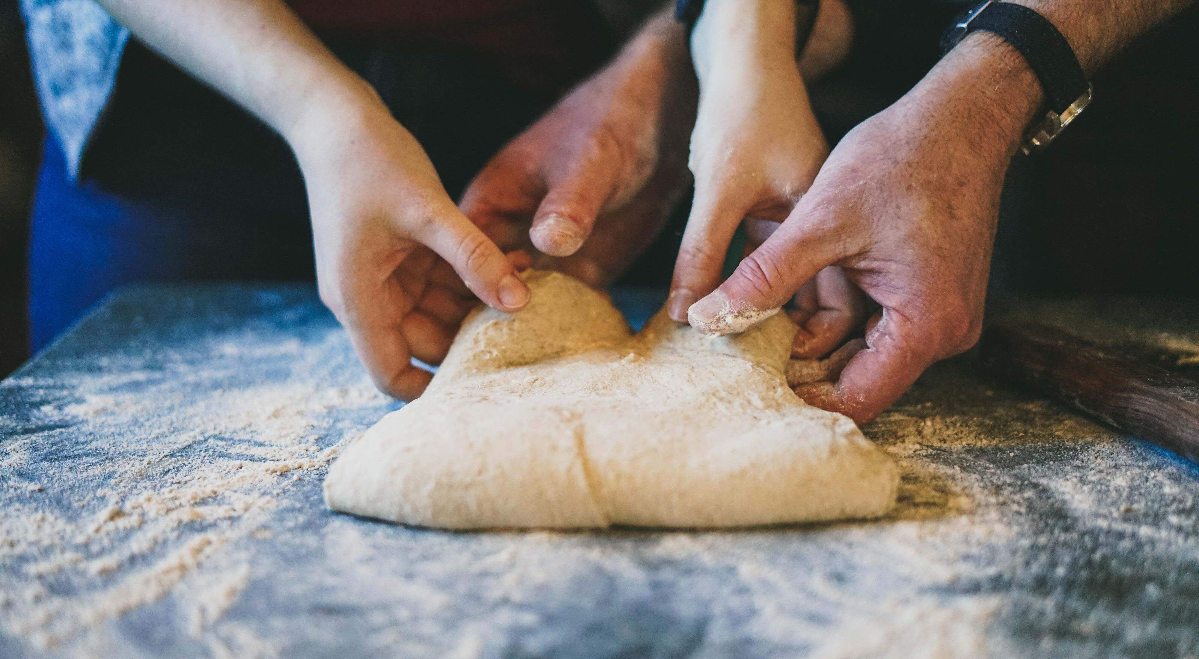 Two sets of hands kneading bread dough on a floured surface.