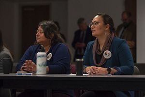 Two women sit at a table. Both have their hands together and resting on the table.
